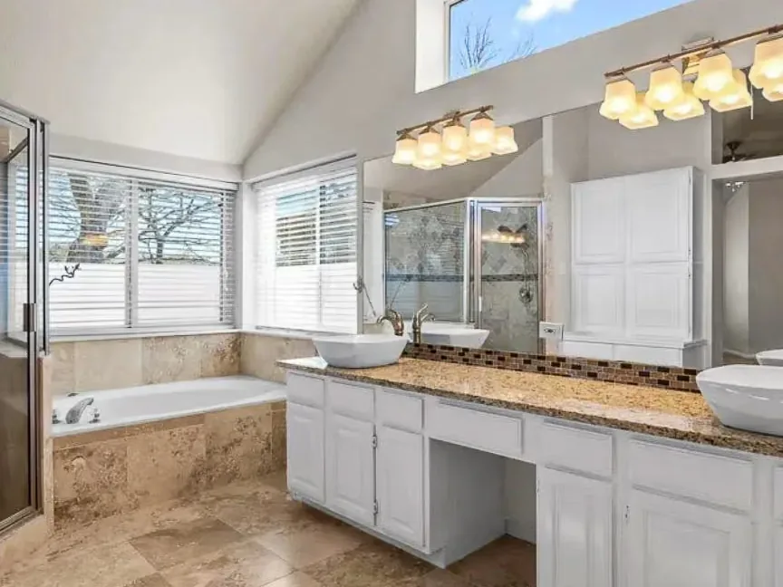 The Oak Tree bathroom before work, with a white vanity, tan granite, a corner tub and travertine tile under a vaulted ceiling.