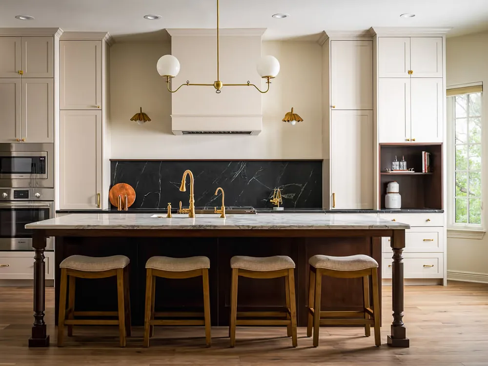 Remodeled Sophora kitchen with cream cabinetry, a dark stone backsplash, brass fittings and a long island with stools.